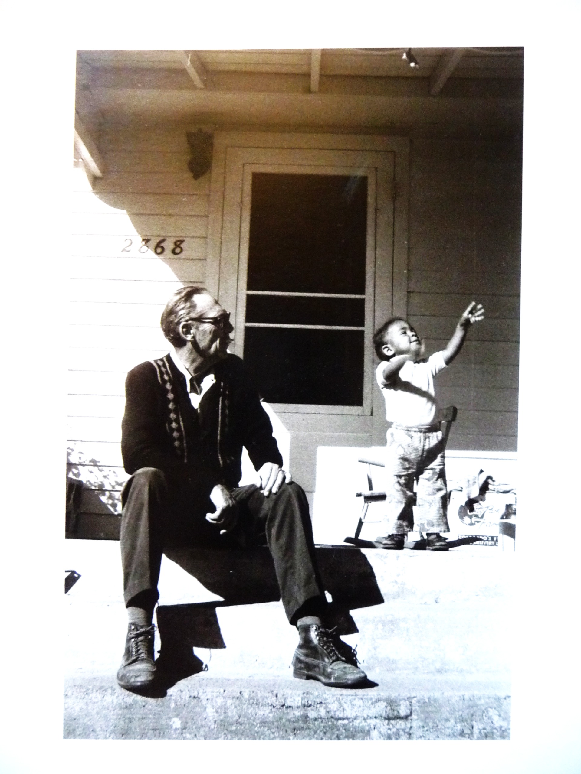 An older white man sits comfortably on a front porch next to a brown-skinned child.  As the man looks on, the child is reaching for the sky.