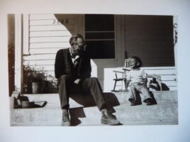 An older white man sits on the porch steps with a brown-skinned child. They appear comfortable and are smiling at each other.